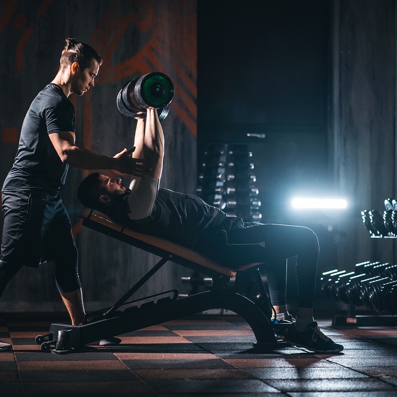 Coach personnel aidant un homme effectuant un développé couché avec haltère sur un banc incliné dans une salle de sport sombre.