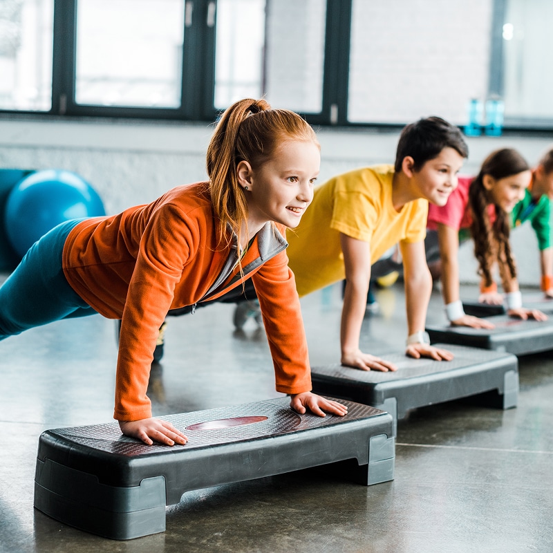 Enfants souriants en position de planche sur des steppers noirs, faisant de l'exercice dans un gymnase lumineux.