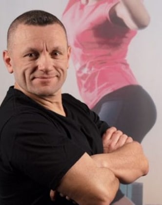 Entraîneur homme souriant, bras croisés, t-shirt noir. En arrière-plan, femme floue en rose avec des fessiers musclés pour le fitness.
