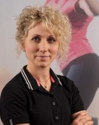 Femme souriante aux cheveux blonds bouclés, portant un polo noir à rayures blanches, sur fond sportif flou. Pose confiante.