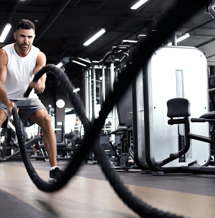 Homme musclé et déterminé s'entraînant avec des cordes de combat dans une salle de sport moderne et sombre. Débardeur blanc.