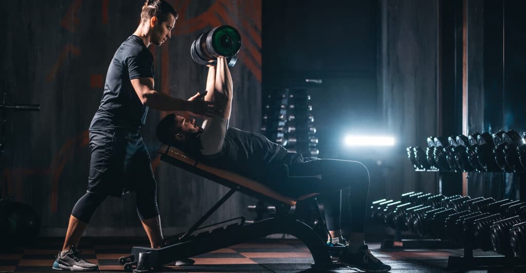 Coach sportif assistant un homme faisant des développés couchés inclinés avec haltères dans une salle de sport sombre.