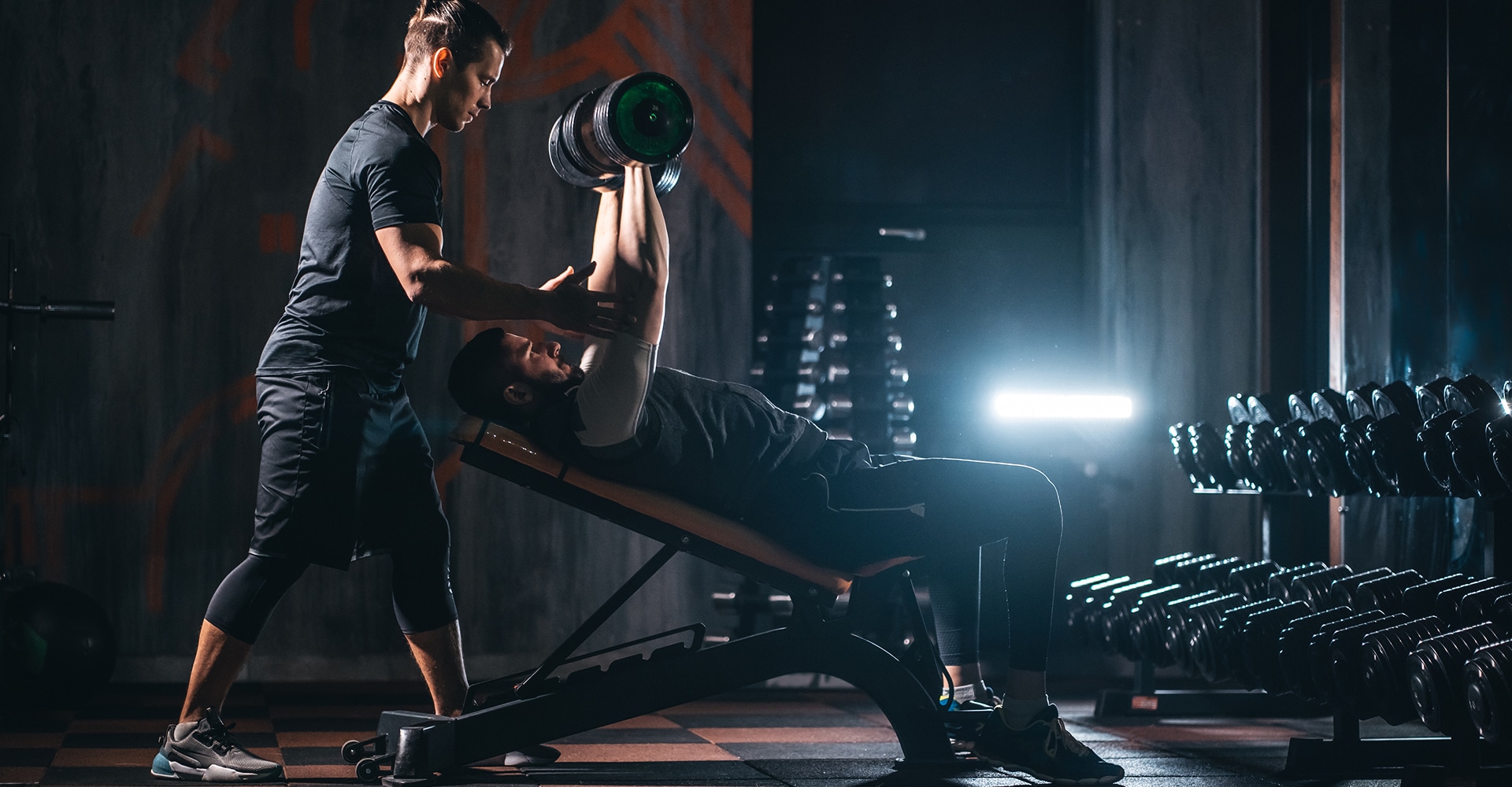 Coach sportif assistant un homme faisant des développés couchés inclinés avec haltères dans une salle de sport sombre.