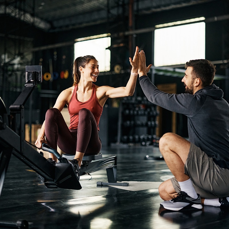 Femme souriante sur rameur et homme accroupi se donnent un high-five dans un club de sport, symbolisant la réussite de l'entraînement.