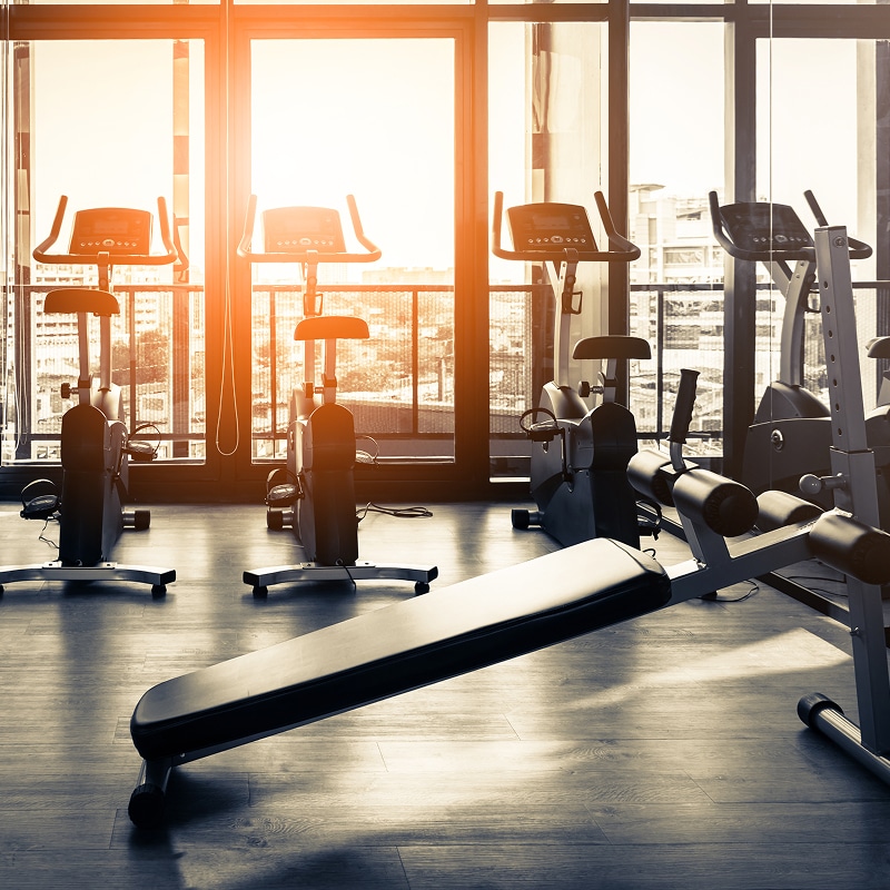 Gym moderne avec vélos d'appartement et banc de musculation. Lumière du soleil orangée à travers les grandes fenêtres donnant sur la ville.