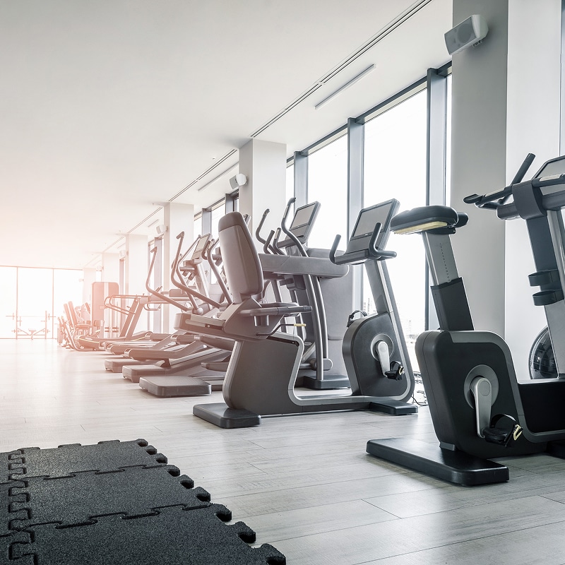 Salle de sport moderne et lumineuse avec rangée de machines de fitness (elliptiques, vélos, tapis) près de fenêtres. Dalles de sol noires.