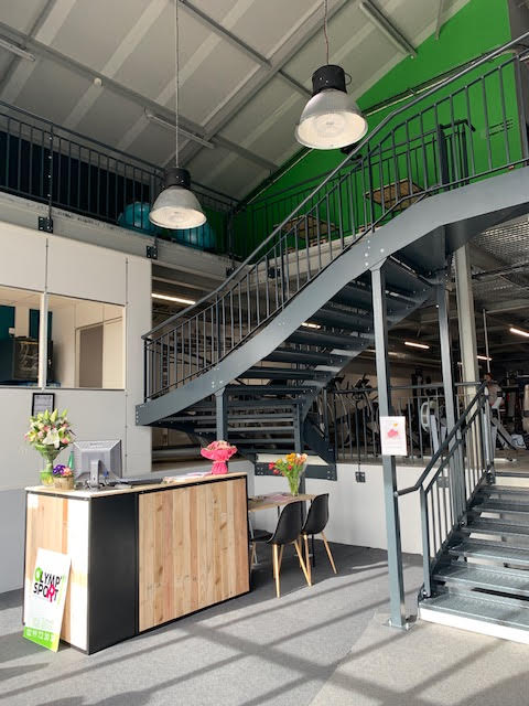 Réception Olymp Sport avec bureau bois et noir. Grand escalier métallique vers salle de sport aux murs verts. Lumières industrielles, signalisation 'OLYMP SPORT'.