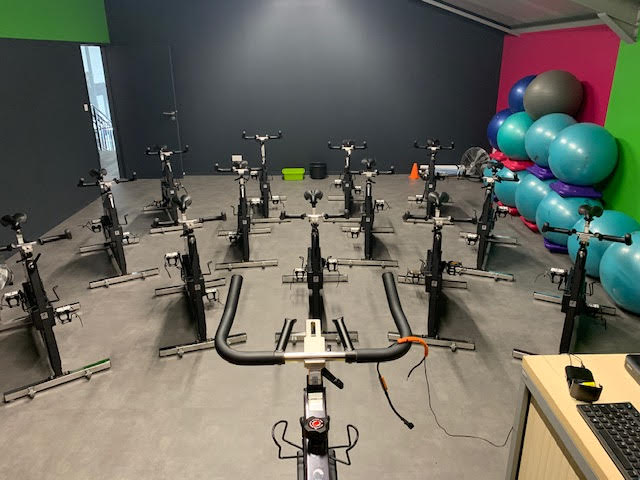 Salle de sport lumineuse avec rangées de vélos de spinning noirs. Ballons de fitness bleus et gris empilés contre un mur coloré.