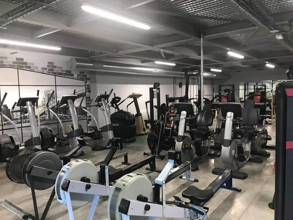 Grande salle de sport moderne avec divers équipements de fitness : rameurs, vélos elliptiques et machines de musculation sous un plafond industriel.