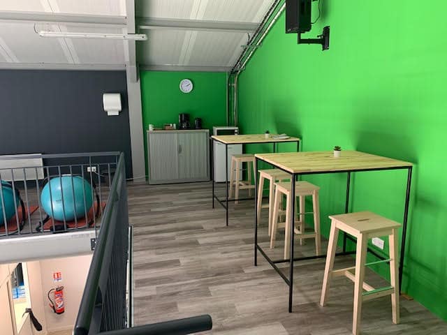Salle de sport moderne avec mezzanine grise, ballons d'exercice bleus et distributeur. Espace détente au mur vert vif, tables hautes en bois et tabourets clairs.